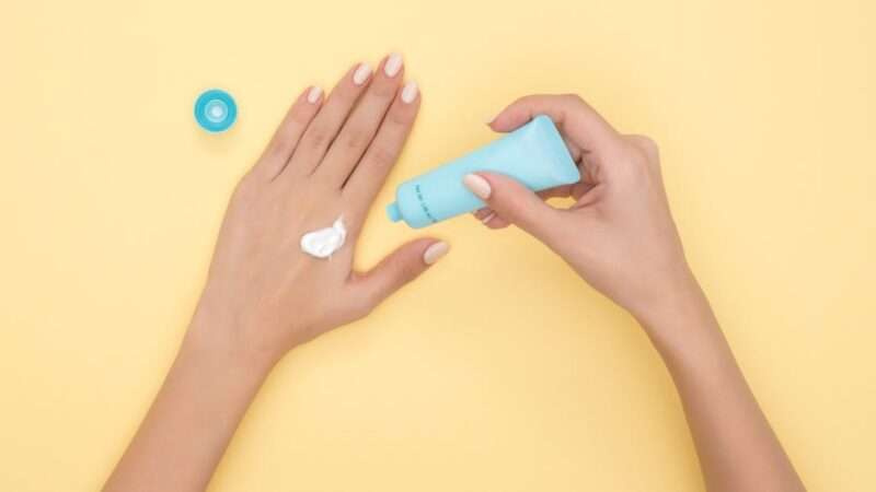 Close-up of a woman's hands applying cream on a pastel yellow background. Ideal for beauty and skincare themes.