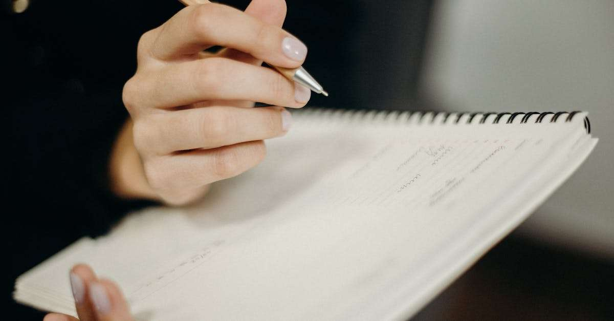 A person's hand holding a pen writing in a spiral notebook, focused on penmanship and note-taking.