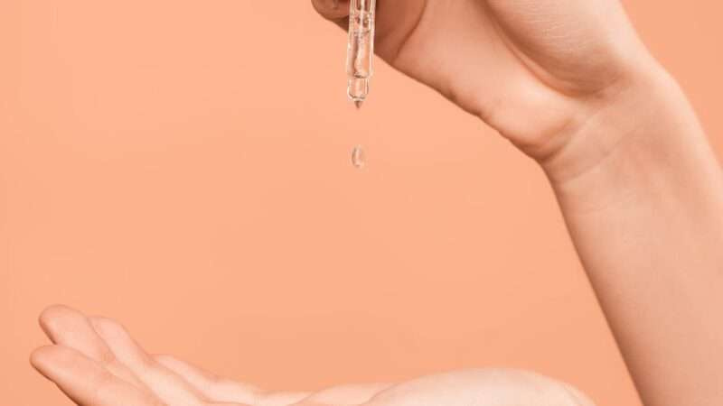 Close-up of hand applying serum with dropper on peach background. Perfect for beauty and skincare routine.