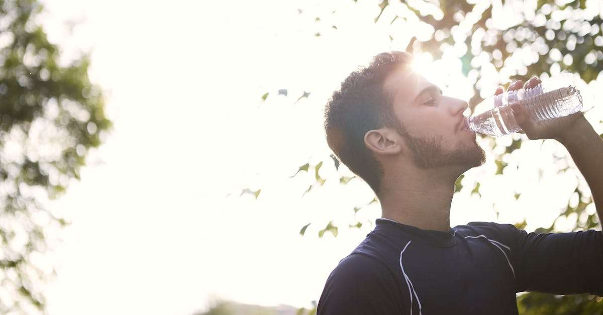 Side view of a young man drinking water from a plastic bottle outdoors in sunlight.