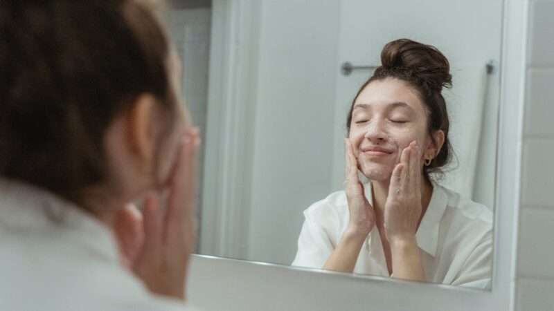 Woman applying soap to her face, reflected in the bathroom mirror, enjoying a skincare routine.